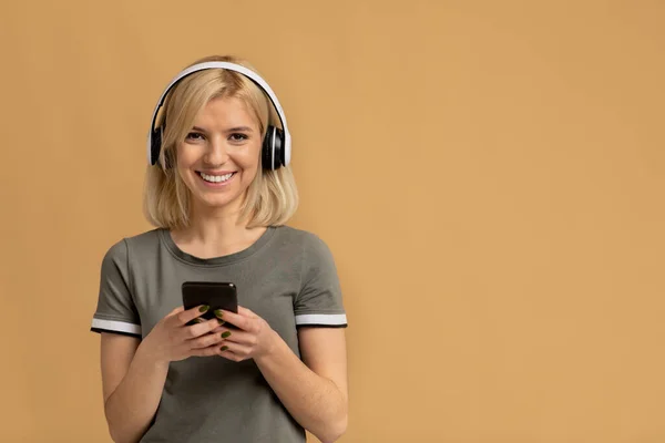 Dispositivos modernos. Mulher feliz usando fones de ouvido sem fio e usando smartphone sobre fundo estúdio bege, espaço de cópia — Fotografia de Stock