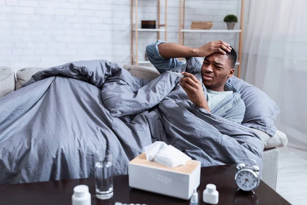Africano teniendo fiebre sosteniendo termómetro acostado en sofá interior — Foto de Stock