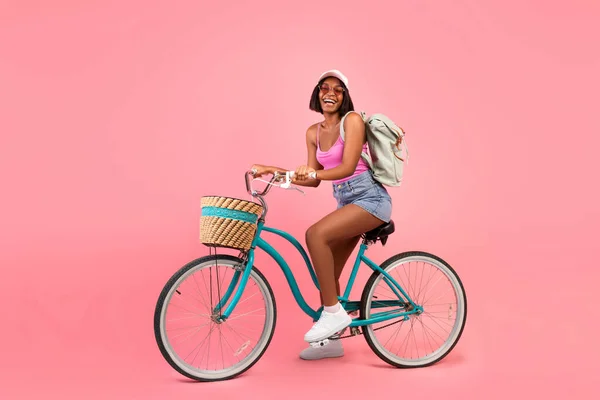 Full length portrait of lovely black woman in casual summer clothes riding bicycle over pink studio background — Stock Photo, Image