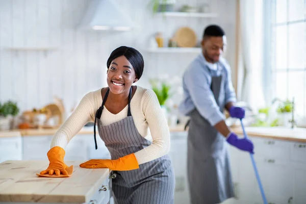 Ritratto di felice signora nera in grembiule e guanti di gomma pulire tavolo da cucina, il suo fidanzato lavaggio pavimento su sfondo — Foto Stock