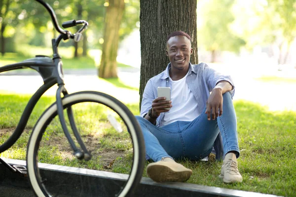 Веселий чорний байкер сидить біля дерева з мобільним телефоном у міському парку — стокове фото