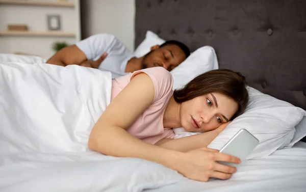 Kíváncsi fiatal nő ellenőrzi az alvó férjét okostelefon az ágyban otthon — Stock Fotó