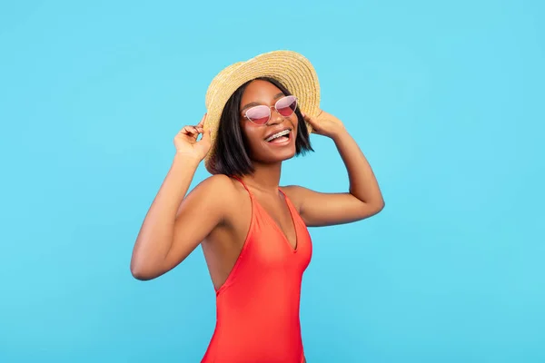 Portrait of lovely black lady in straw hat and swimsuit posing and smiling at camera on blue background — Stock Photo, Image