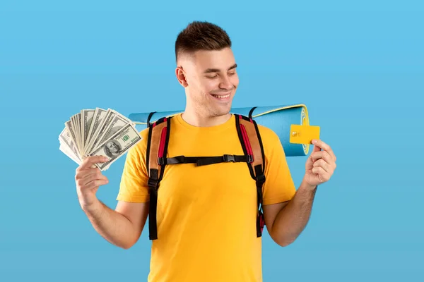 Presupuesto de viaje. Retrato de un joven guay con mochila, tarjeta de crédito y dinero estadounidense sobre fondo de estudio azul —  Fotos de Stock