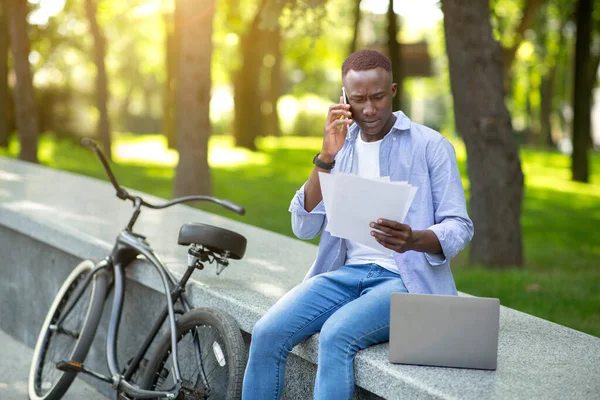 Homem negro preocupado com documentos falando no smartphone perto de sua bicicleta no parque da cidade — Fotografia de Stock