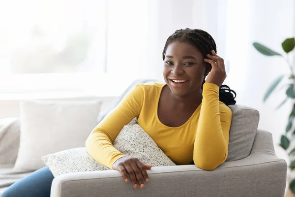 Positiva dama afroamericana posando en el interior del hogar, sentada en un sofá cómodo —  Fotos de Stock