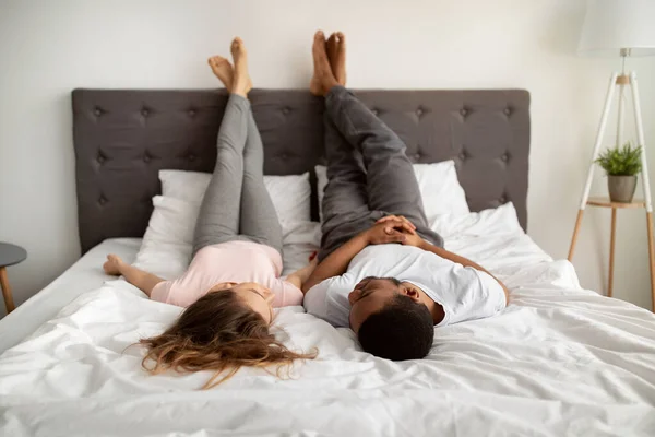 Millénaire interracial couple dans l'amour couché sur le lit à la maison, levant les jambes, se parler — Photo