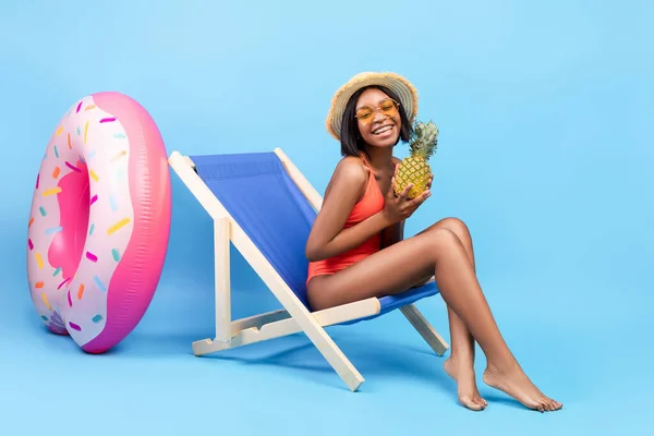 Vacaciones de verano. Mujer joven en bikini sosteniendo piña, sentada en sillón y sonriendo sobre fondo azul — Foto de Stock