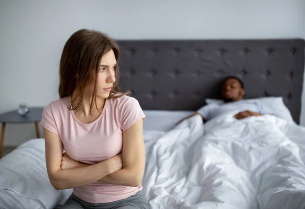 Молодая межрасовая пара переживает кризис отношений, депрессивная женщина сидит на кровати, ее муж спит, пустое пространство — стоковое фото
