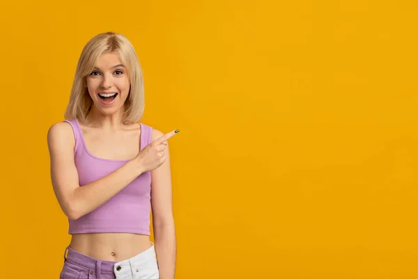 Για δες αυτό. Χαρούμενη ξανθιά γυναίκα που δείχνει με το δάχτυλο στον κενό χώρο, χαμογελώντας στην κάμερα πάνω από κίτρινο φόντο — Φωτογραφία Αρχείου