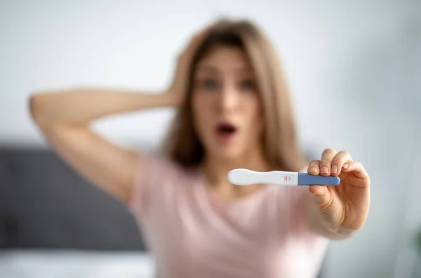 Mujer joven conmocionada mostrando prueba de embarazo positiva en la cámara, agarrando su cabeza aterrorizada, en interiores —  Fotos de Stock