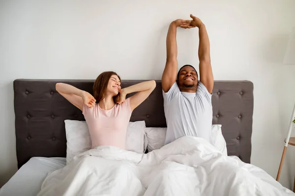 Fiatal multiracionális pár nyújtózkodik az ágyon, miután felébredtek otthon. — Stock Fotó