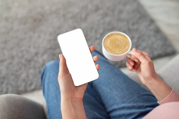 Female hands holding smartphone with blank screen and cup of coffee, sitting on sofa at home