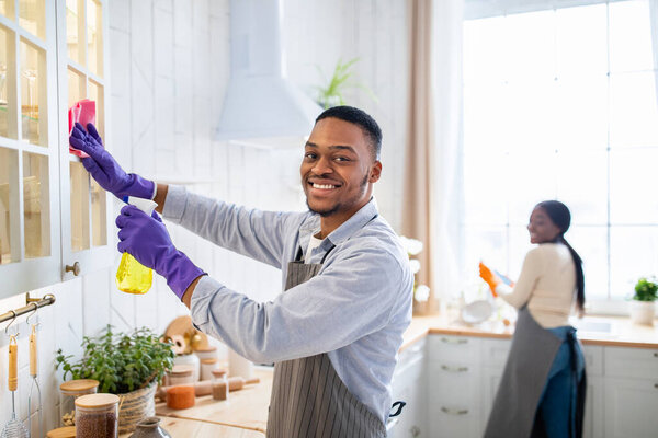 Attractive African American guy cleaning kitchen cabinet with detergent, his wife helping him on background, copy space