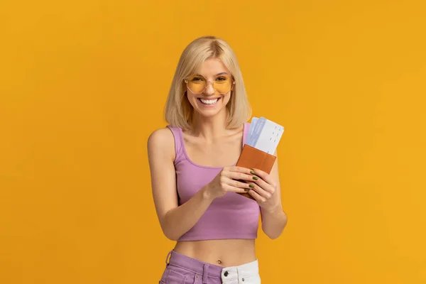 Καλοκαιρινό. Ευτυχισμένη κυρία με πολύχρωμα γυαλιά ηλίου κρατώντας διαβατήριο και εισιτήρια, στέκεται πάνω από το κίτρινο φόντο — Φωτογραφία Αρχείου