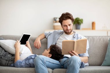 Farklı ilgi alanları. Genç babası kağıt kitap okurken küçük oğlu dijital tablet kullanıyor, birlikte kanepede dinleniyorlar.