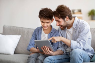 Genç bir baba oğluna dijital tablet üzerinde yeni uygulamalar kullanmayı öğretiyor, birlikte zaman geçirirken internette sörf yapıyor.