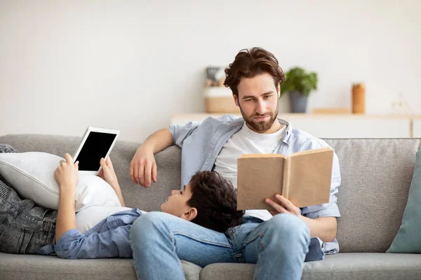 Andere interesses. Jonge vader leest papieren boek terwijl zijn tiener zoon met behulp van digitale tablet, rusten samen op de bank — Stockfoto