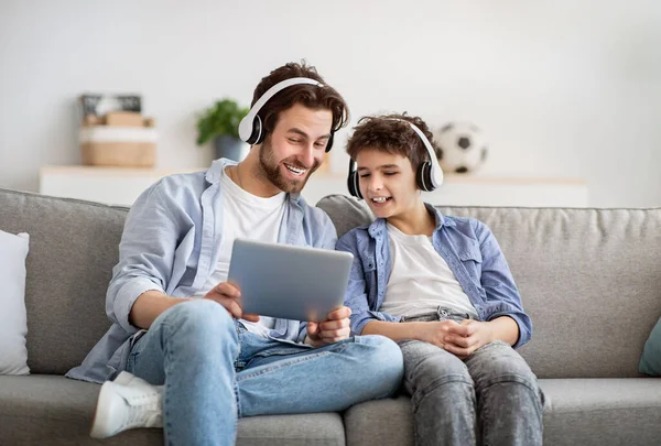 Père et fils modernes regardant des vidéos sur tablette numérique, portant des écouteurs sans fil et souriant, surfant sur Internet — Photo