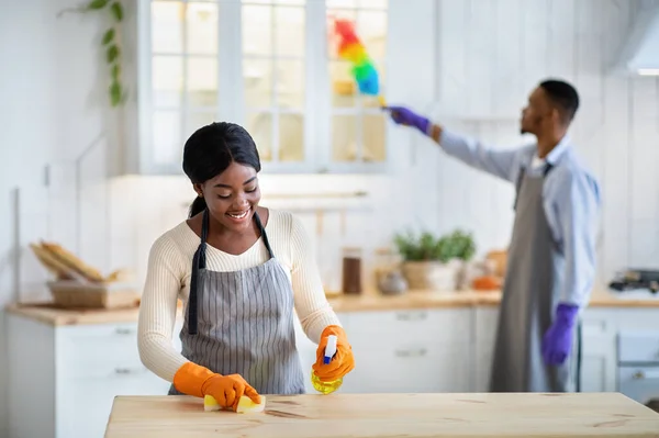 Schöne schwarze Dame putzt Küchentisch mit Spray, während ihr Mann Staub auf Hintergrund, freier Raum — Stockfoto