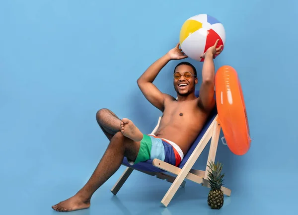 Longitud completa del chico negro guapo en traje de baño sentado en silla de salón, sosteniendo la bola inflable sobre fondo azul —  Fotos de Stock