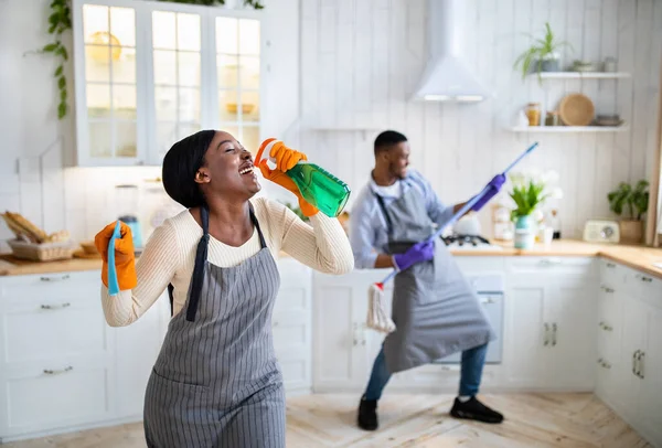 快乐的黑人女士用洗涤剂作为话筒，她的男朋友在厨房里像吉他一样弹奏拖把，复制空间 — 图库照片