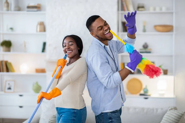 快乐的黑人男人和他的女朋友在打扫房间的时候唱歌，用拖把和粉碎机当麦克风 — 图库照片