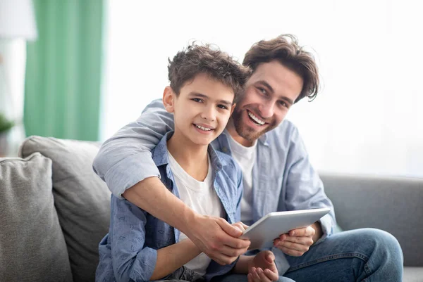 Семейное время. Счастливые папа и сын, пользуясь цифровым планшетом, просматривая интернет дома, сидя вместе на удобном диване — стоковое фото