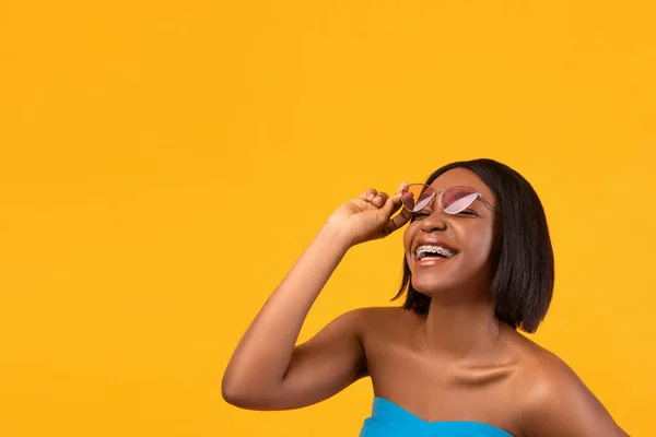 Portrait of happy black lady in dental braces touching sunglasses and smiling on orange background, copy space — Stock Photo, Image