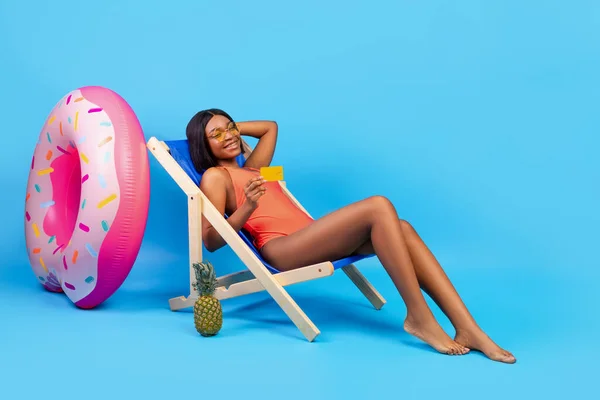 Orçamento de férias de verão. Mulher preta bonita em suimsuit relaxante na cadeira lounge com cartão de crédito no fundo azul — Fotografia de Stock