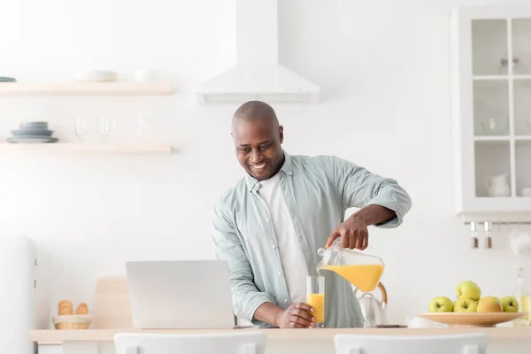 Morning routine. Happy mature black man having video chat with family on laptop computer, pouring juice in kitchen — Stock Photo, Image