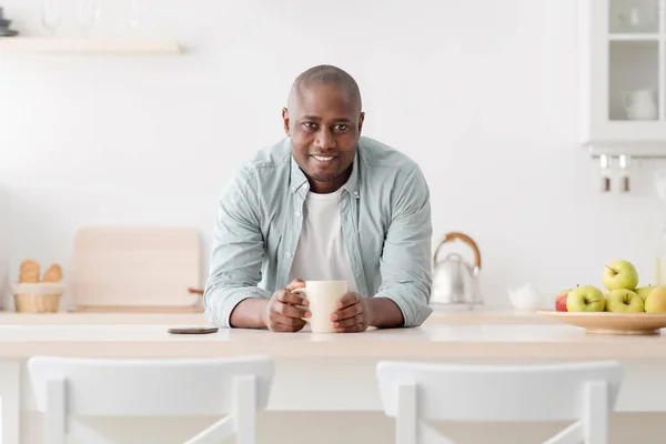 Harmonie života. Šťastný afroameričan starší muž pije kávu doma v kuchyni, dívá se na fotoaparát a usmívá — Stock fotografie