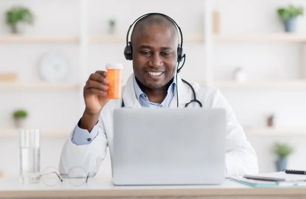 De dokter beveelt medicijnen aan. Afro-Amerikaanse therapeut in hoofdtelefoon tonen pot pillen naar laptop scherm — Stockfoto