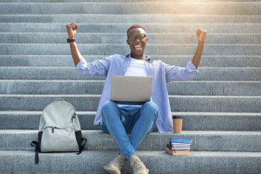 Heyecanlı Afrikalı Amerikalı öğrenci şehir merkezindeki merdivenlerde dizüstü bilgisayarın önünde EVET işareti yapıyor.