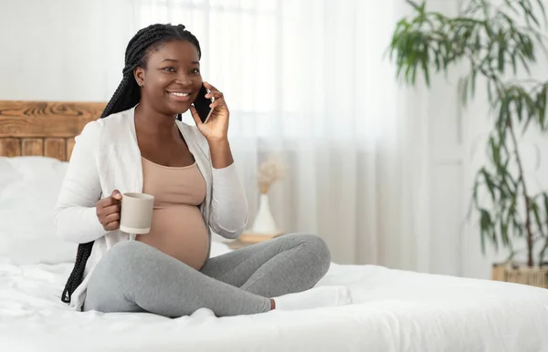 Hamile eğlencesi. Gülümseyen siyahi anne cep telefonuyla konuşuyor ve çay içiyor. — Stok fotoğraf