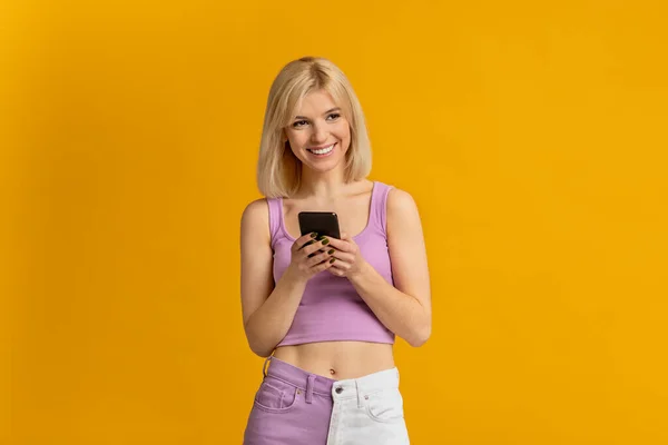 Ενθουσιασμένη κυρία με smartphone που χρησιμοποιεί νέα εφαρμογή κινητού τηλεφώνου πάνω από κίτρινο φόντο στούντιο — Φωτογραφία Αρχείου