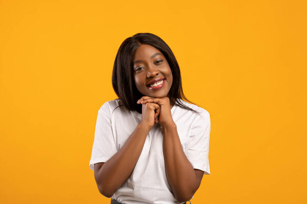 Sweet black lady showing tenderness on yellow studio background, touching her chin with both things and smiling at camera, african american woman flirting or seeing something heart melting