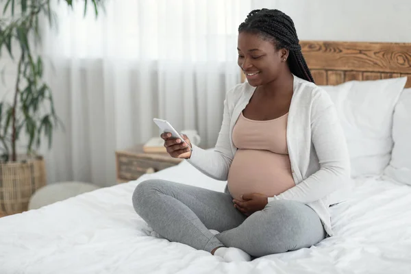 Счастливая молодая афроамериканская беременная женщина расслабляется со смартфоном дома — стоковое фото