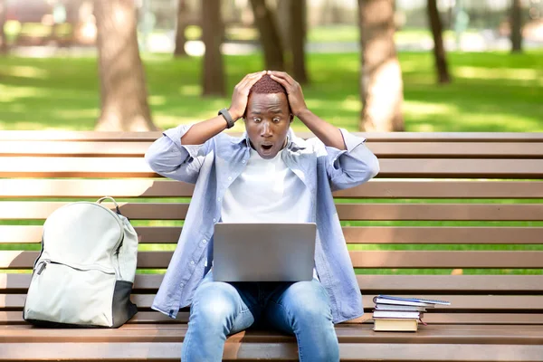 Terrorizzato uomo nero guardando lo schermo del computer portatile e afferrando la testa nel panico sulla panchina al parco — Foto Stock