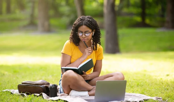 Удумлива студентка з ноутбуком і блокнотом вивчає онлайн в парку — стокове фото