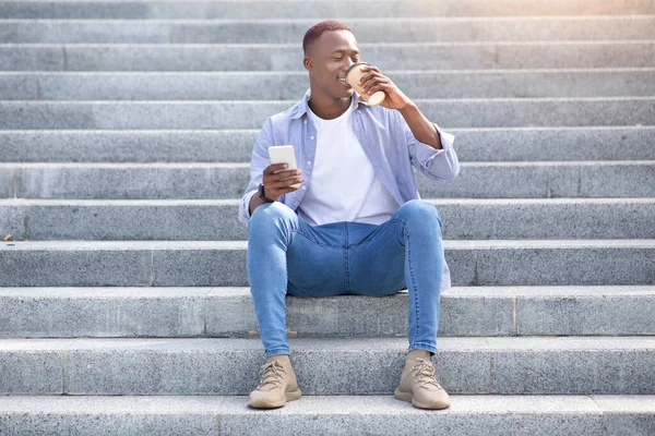 外の階段に座っている間にテイクアウトコーヒーを飲み、携帯電話を使用してハンサムな黒人男性 — ストック写真
