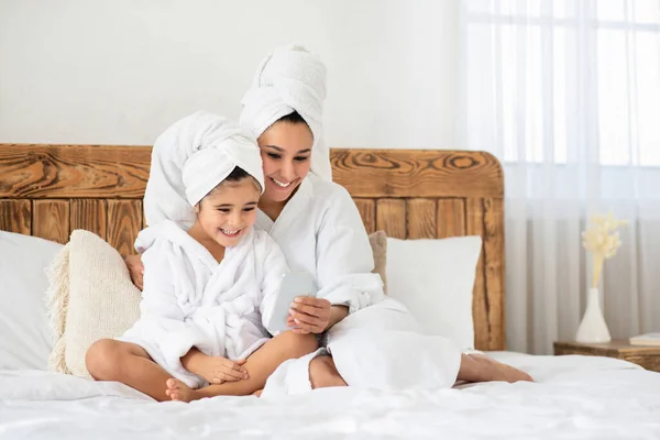 Sorrindo mãe e menina sentados na cama, usando smartphone — Fotografia de Stock