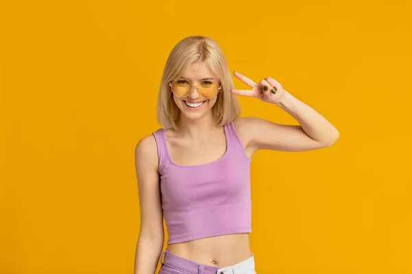 Mujer alegre con gafas de sol con estilo y mostrando la paz v-signo, posando sobre fondo de estudio amarillo —  Fotos de Stock