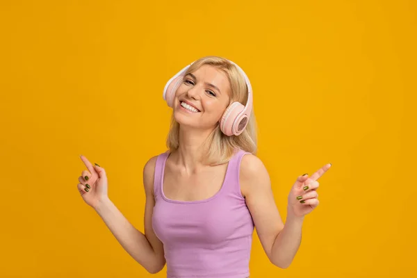 Carefree žena ve sluchátkách dacing na její oblíbenou hudbu a úsměv na žlutém pozadí studia — Stock fotografie