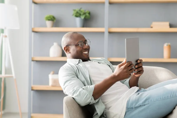 Afrikanischer Mann liest E-Book auf digitalem Tablet oder surft im Internet, genießt Wochenende und ruht sich im Sessel aus — Stockfoto