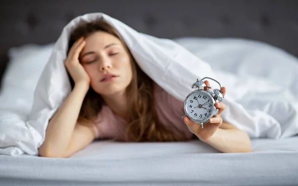 Ospalá mladá dáma ležící pod přikrývkou v posteli s budíkem, ráno se špatně probouzí — Stock fotografie