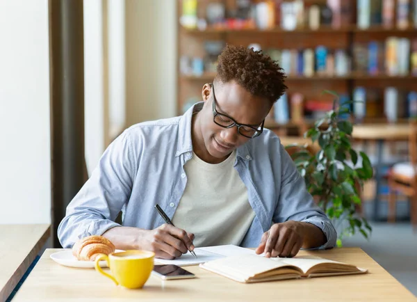 Hombre negro trabajador escribiendo información de libro de texto, preparándose para la prueba en la cafetería —  Fotos de Stock