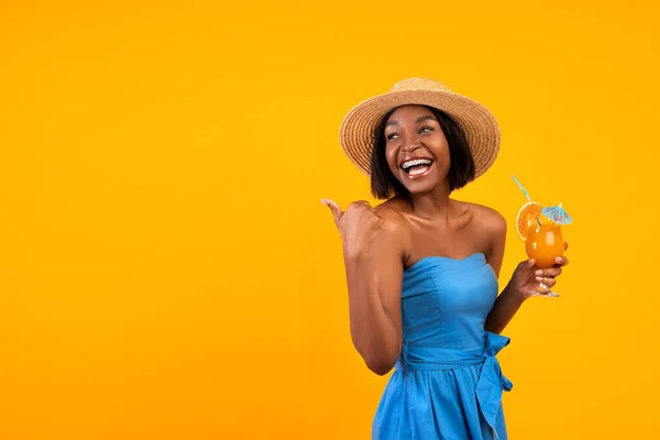 Yazlık elbiseli ve şapkalı mutlu siyah kadın ferahlatıcı bir kokteyl içiyor, turuncu arka plandaki boş alanı işaret ediyor. — Stok fotoğraf
