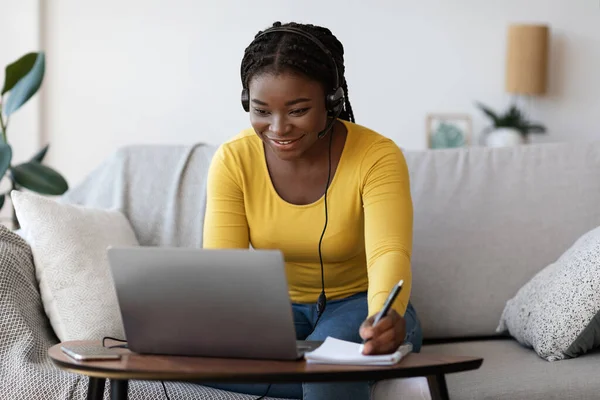 Glimlachende zwarte dame in headset schrijven notities tijdens het kijken webinar op laptop — Stockfoto