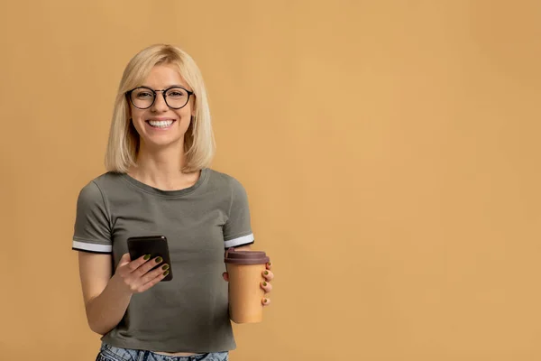 Modern student som håller hämtmat kaffe och skriva på smartphone på beige bakgrund med tomt utrymme — Stockfoto